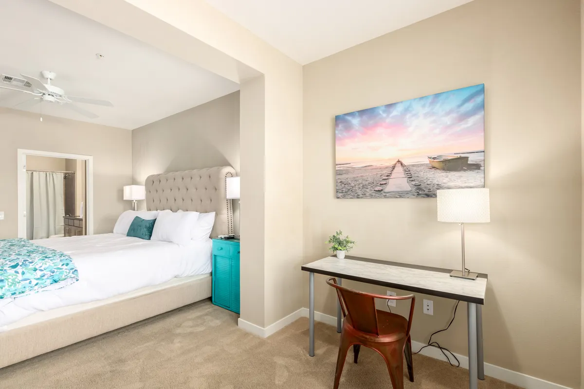 A tranquil bedroom scene with a bed outfitted in decorative pillows, a turquoise nightstand, and calming beach-themed wall art. A desk with a lamp sits adjacent to the window, creating a dedicated office or sitting nook ideal for work or peaceful reflection.