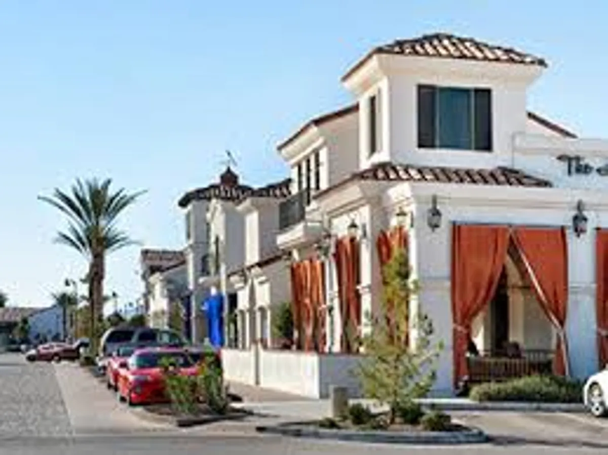 A bustling street scene with decorative curtains, palm trees, colorful cars, and architectural details. People are seen walking, socializing, shopping, and enjoying the lively neighborhood vibe, indicative of community events or outdoor markets.