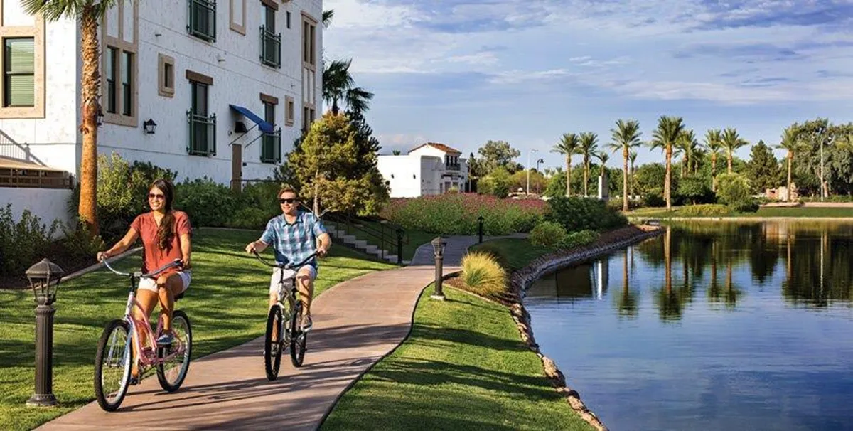 This image showcases a vibrant outdoor scene with bicyclists riding along a lakeside pathway lined with palm trees. The sparkling water and lush greenery evoke a sense of leisure and activity, while the bright, sunny weather adds to the cheerful atmosphere.