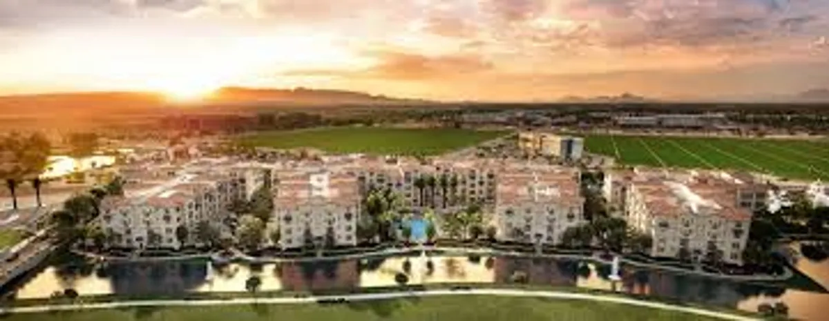 This image captures modern apartment buildings set beside a reflective water surface, surrounded by lush greenery and highlighted by a warm sunset sky. The atmosphere is tranquil and inviting, with visual cues of outdoor relaxation and gatherings near the water's edge.