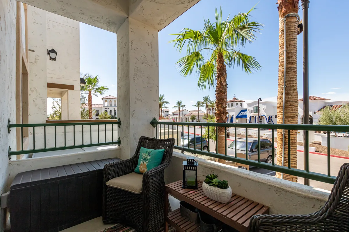 A cozy, inviting balcony scene with palm trees in the background, comfortable outdoor seating with decorative cushions, a lantern for ambiance, and a planter adding a touch of greenery. The lighting captures the morning sunshine filtering through the leaves, suggesting a perfect spot for relaxing, reading, or enjoying a coffee.