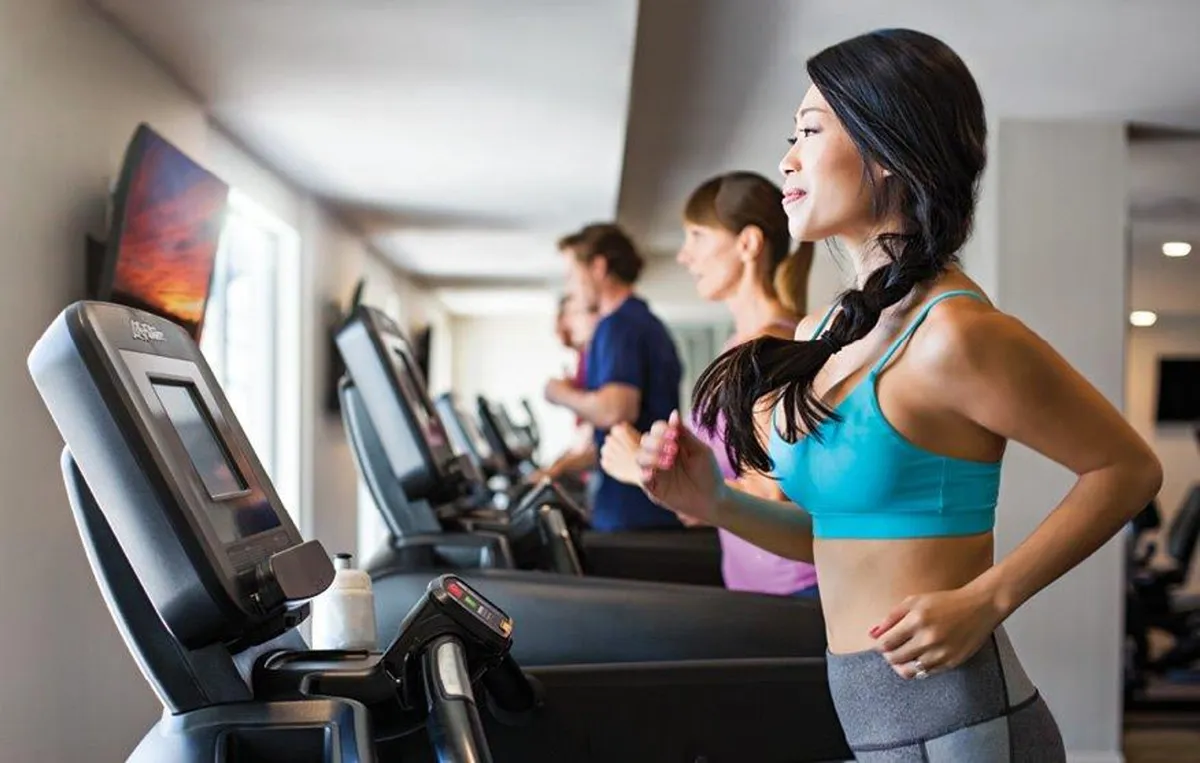 A bright, modern fitness center interior with several treadmills in use, people actively exercising, and a large television screen displaying fitness content. The space is energetic and contemporary, designed to motivate residents to focus on their health goals.