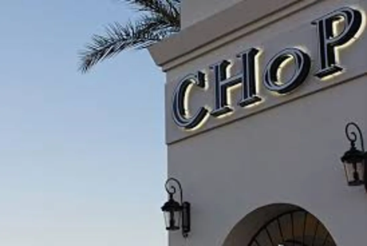 The image features a warmly lit restaurant with a prominent sign, decorative lanterns, and a palm tree, suggesting a lively yet welcoming dining environment. The ambiance hints at evening social gatherings and culinary delights in an outdoor setting.