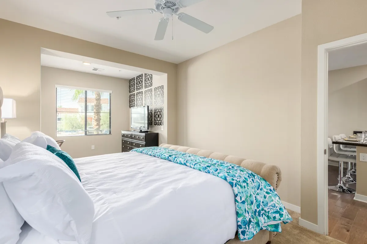 A serene bedroom featuring a luxurious king-sized bed adorned with a decorative throw, elegant wall art above the headboard, and a large window flooding the space with natural light. The setup is uncluttered and designed for comfort, evoking the feel of a high-end retreat.