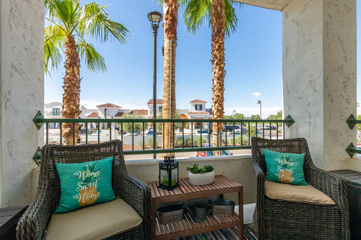 A north-facing balcony furnished with wicker chairs and decorative cushions, palm trees in the background, and a view overlooking the parking lot. The space is styled for comfort and casual outdoor enjoyment, with a small table and shaded ambiance, suggesting suitability for morning coffee or afternoon relaxation.