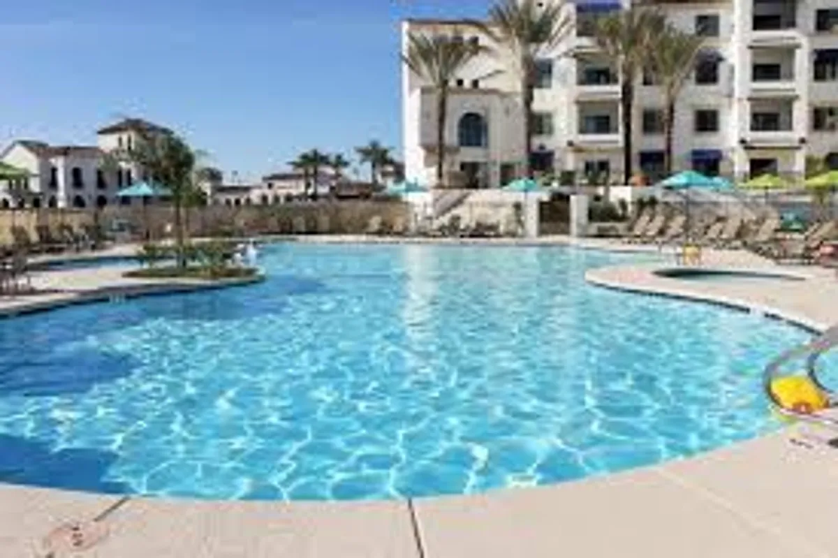 This lively exterior image showcases a sparkling swimming pool surrounded by comfortable lounge chairs and lush palm trees, with modern community buildings in the background. The inviting pool setting is bathed in warm sunlight, suggesting a perfect spot for relaxation, swimming laps, or socializing with others.