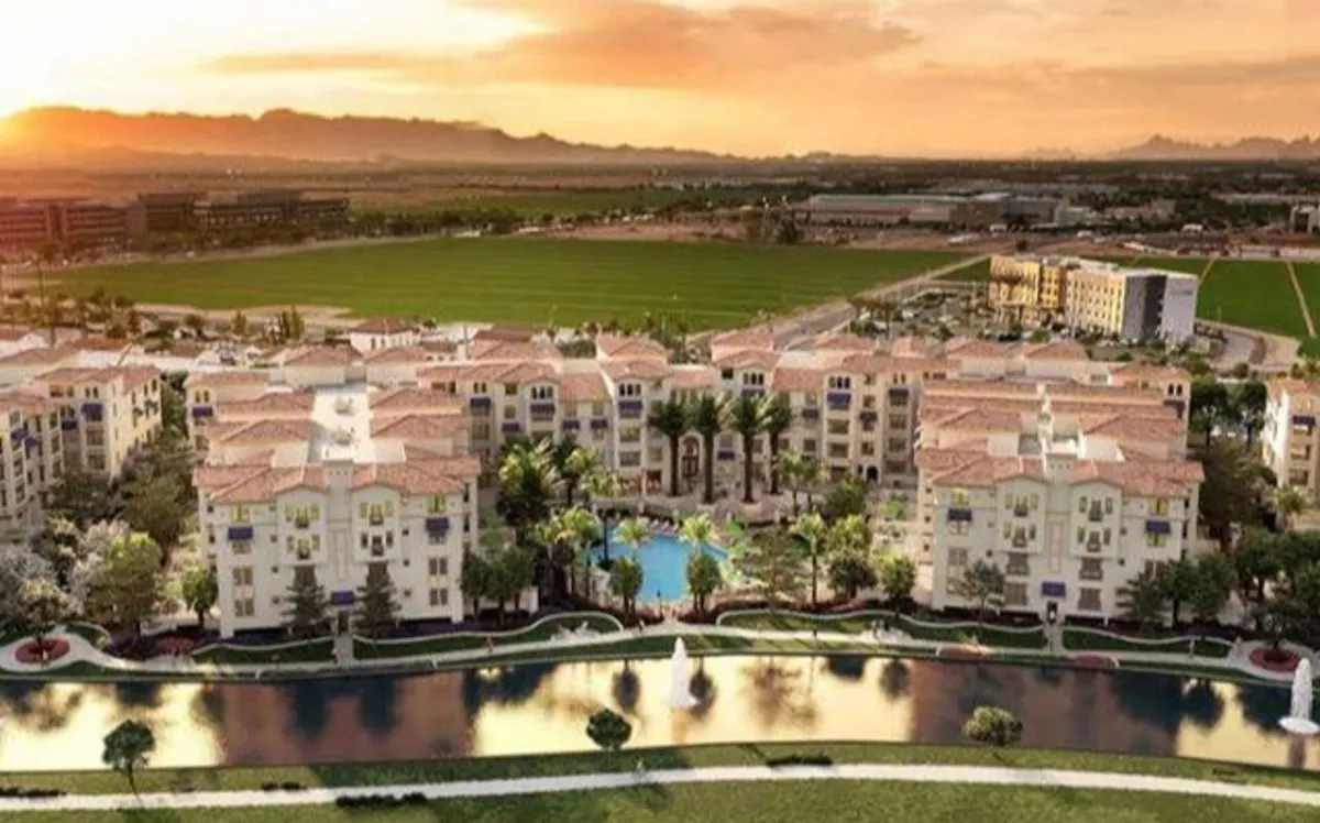 A luxurious outdoor pool area surrounded by palm trees and landscaped greenery, with modern apartment buildings as a backdrop. Water features enhance the tranquil resort vibe, and there are inviting places to relax by the pool or walk along the water's edge.